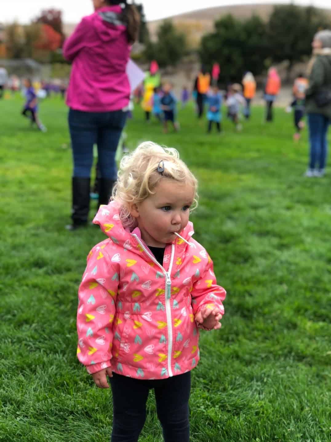 girl in a pink raincoat eating a lollipop while standing in a grassy field