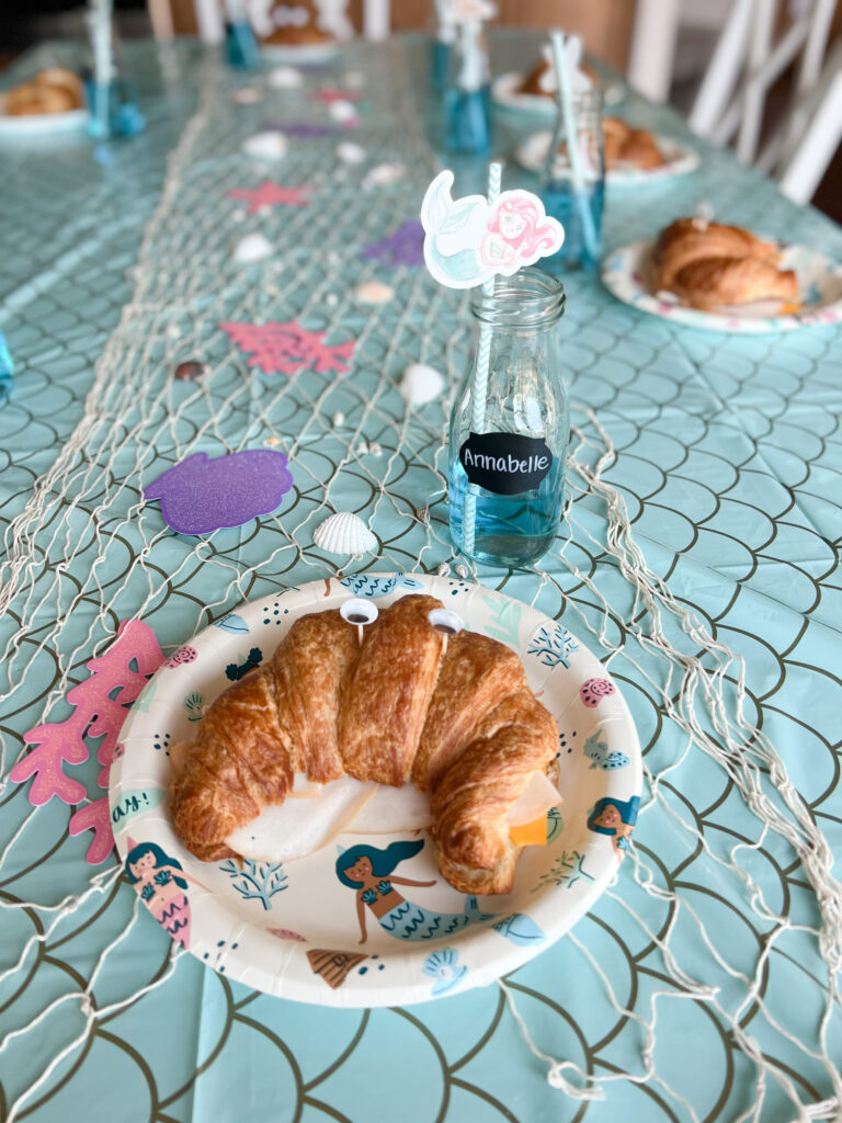croissant sandwich with googly eyes stuck into to make it look like a crab. Blue drink on a blue tabelcloth.